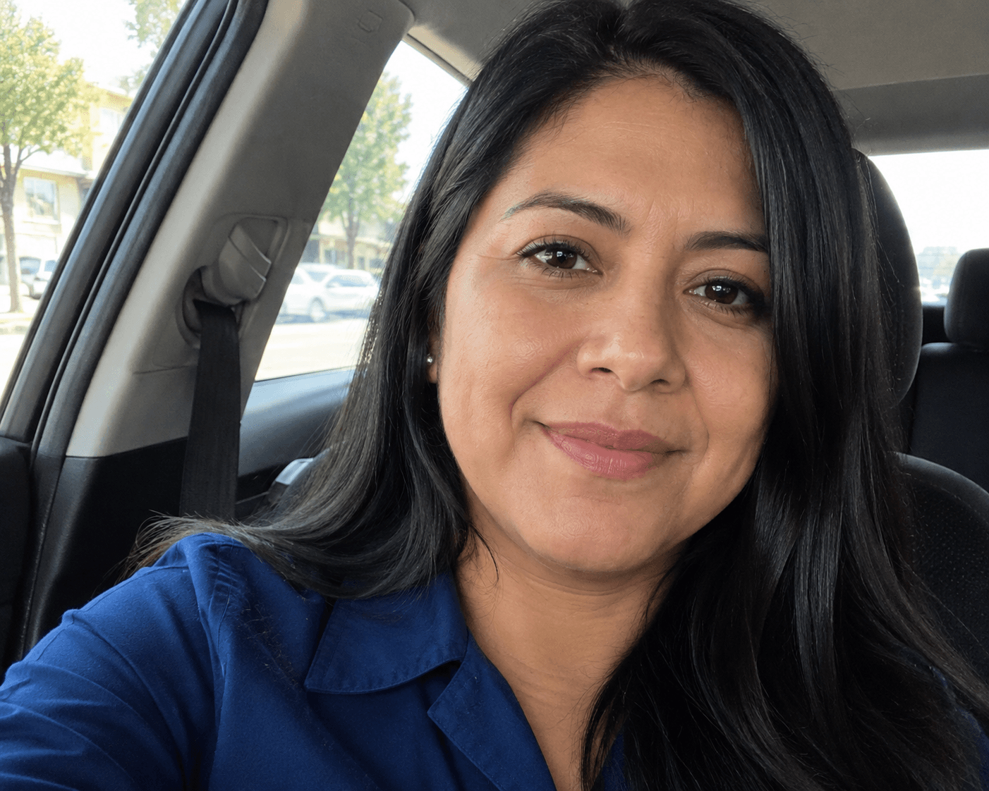 Smiling woman with long black hair wearing a blue shirt sitting in a car.